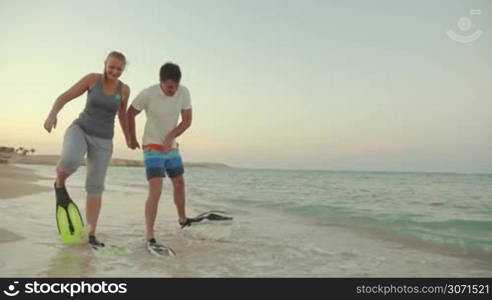 Slow motion steadicam shot of young man and woman walking in flippers along the sea shore. They look funny and clumsy