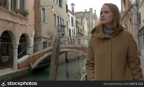 Slow motion steadicam shot of young woman walking on sidewalk along the canal in old city of Venice. She drinking coffee and looking upon ancient architecture