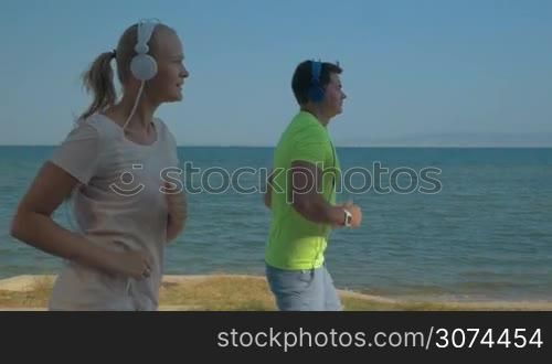 Slow motion steadicam shot young man and woman having sports training at the seaside. They running along the coast and listening to music in headphones. Healthy and active lifestyle