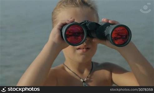 Slow motion view of small boy watching with binoculars with red glasses on landscape against blurred sea