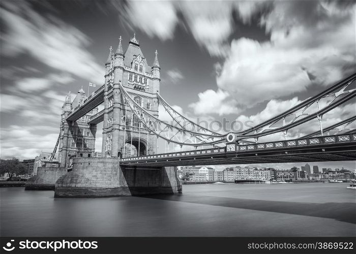 Slow shutter black &amp; white image of Tower Bridge over a blurred river Thames in London with flags flying, clear skies and fluffy clouds