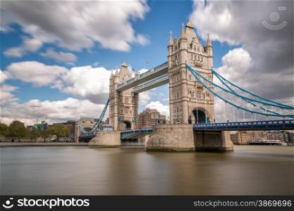 Slow shutter image of Tower Bridge over a blurred river Thames in London with flags flying, blue skies and fluffy clouds