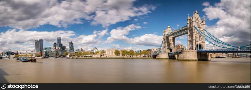Slow shutter panorama of Tower Bridge, the Tower of London and the city with a blurred river Thames in foreground with blue skies and fluffy clouds
