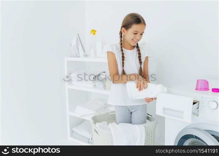 Small adorable busy girl stands in laundry basket, pours detergent in washing machine compartment, has glad expression, long hair combed in pigtails, does housework in laundry room. Cleanliness