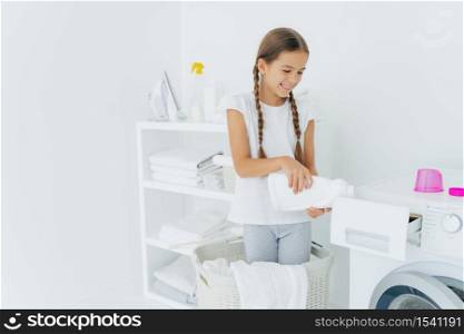 Small adorable busy girl stands in laundry basket, pours detergent in washing machine compartment, has glad expression, long hair combed in pigtails, does housework in laundry room. Cleanliness