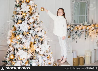 Small adorable female child in white sweater and trousers holds toy for decoration, decorates New Year tree. Cheerful little kid near Christmas tree. New Year s Eve. Holidays and people concept