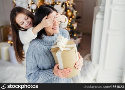 Small adorable female kid prepares surprise for her mother, closes eyes and gives wrapped present as stand at New Year background. Pretty female recieves gift from daughter. Surprisment concept
