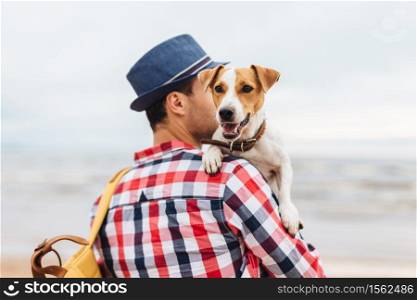 Small beautiful dog on owner`s hands. Stylish young male model dressed in fashionable clothes carries his pet and bag, stand at seashore, come to admire wonderful landscapes and breath fresh air