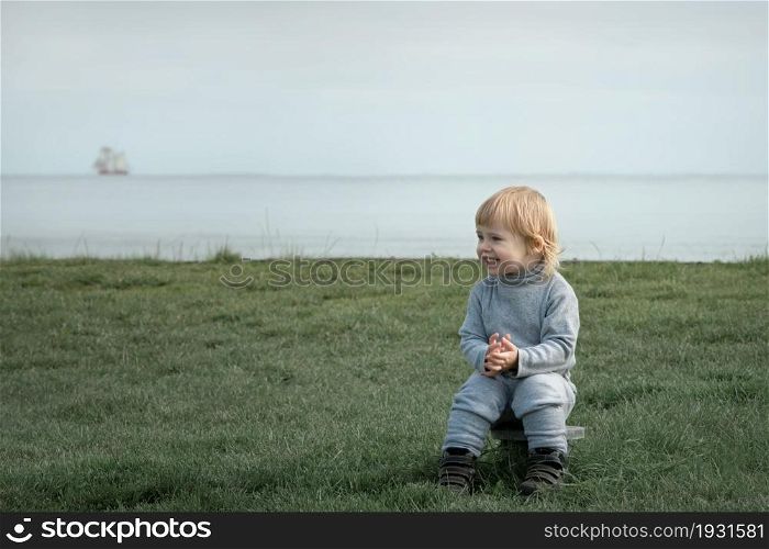 Small blond child sits on grass and smiles on the seashore