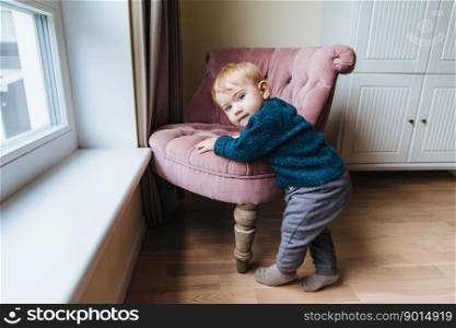 Small blonde child leans at armchair, plays alone at home. Little infant teaches to go. Adorable kid stands near chair, going to play mischievous tricks. Chilren, childhood and lifestyle concept