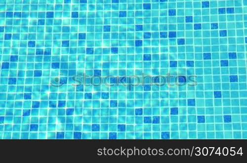 Small blue tiles on the floor of swimming pool, there are sun flecks on the water.