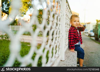 Small boy in red shirt sitting on the fence by the house looking to the street