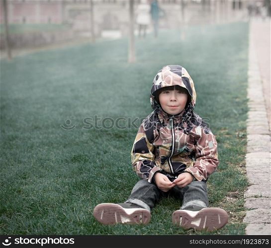 Small boy is in brown jacket sits on grass in the park, fog background, fall season
