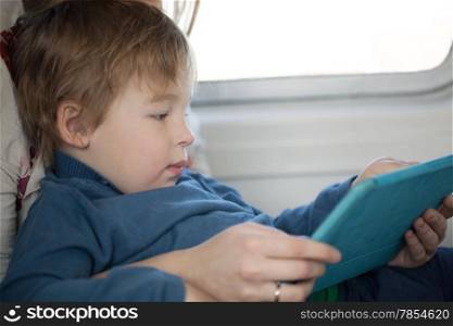 Small boy sitting on his mothers lap alongside a window looking at a tablet in an airplane as he travels on vacation, side view candid portrait