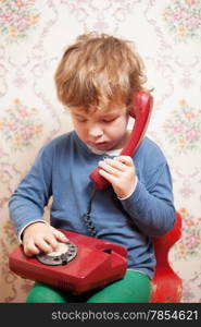 Small boy talking on a red telephone sitting on a plastic chair holding the instrument on his lap