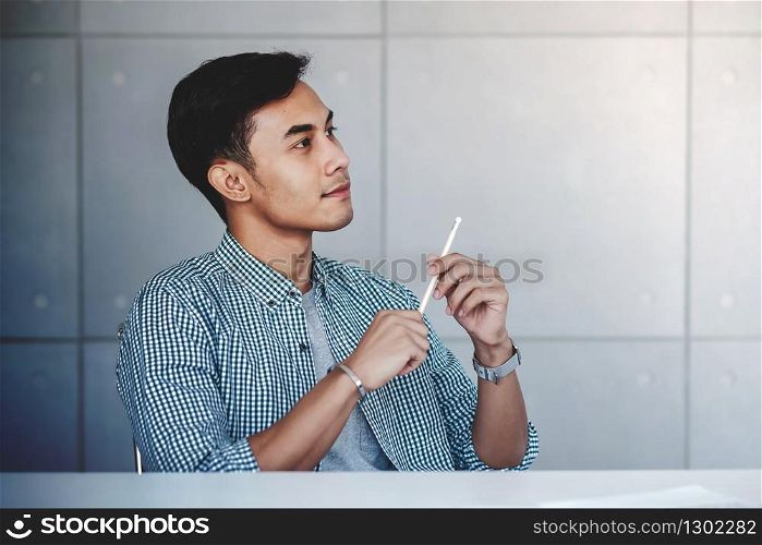 Small Business and Own Proprietor Concept. Young Asian Businessman Sitting at Modern Loft Workplace in Casual Dress. Smiling with Thoughtful Face and looking away