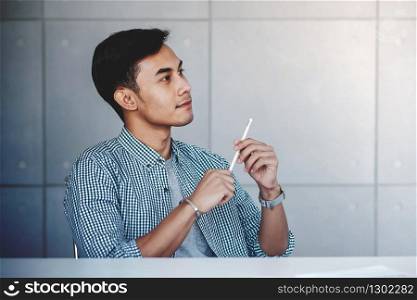 Small Business and Own Proprietor Concept. Young Asian Businessman Sitting at Modern Loft Workplace in Casual Dress. Smiling with Thoughtful Face and looking away