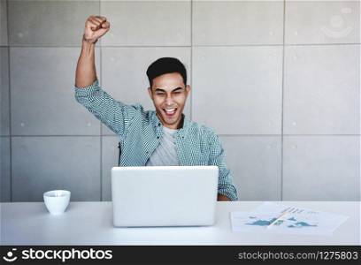 Small Business and Successful Concept. Young Asian Businessman Glad to recieve a Good News or High Profits from Computer Laptop, Own Business Achieves Goals. Raising Arms to Celebrating Success