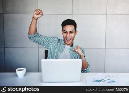 Small Business and Successful Concept. Young Asian Businessman Glad to recieve a Good News or High Profits from Computer Laptop, Own Business Achieves Goals. Raising Arms to Celebrating Success