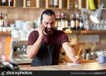 small business, communication, people and service concept - happy man or waiter in apron calling on smartphone at bar or coffee shop. happy man or waiter at bar calling on smartphone