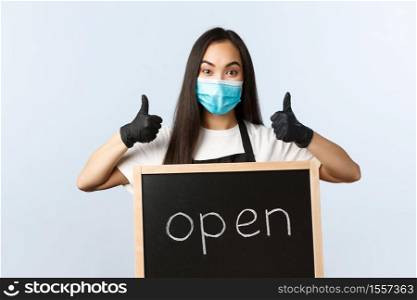 Small business, covid-19 pandemic, preventing virus and employees concept. Upbeat asian female store worker, cafe staff or barista in medical mask show thumb-up and we are open sign.. Small business, covid-19 pandemic, preventing virus and employees concept. Upbeat asian female store worker, cafe staff or barista in medical mask show thumb-up and we are open sign
