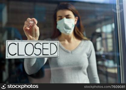 small business, pandemic and service concept - young woman in protective medical face mask hanging banner with closed word on door or window. woman in mask hanging banner closed on door