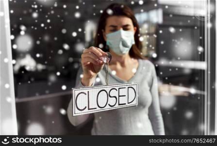 small business, pandemic and service concept - young woman in protective medical face mask hanging banner with closed word on door or window in winter over snow. woman in mask hanging banner closed on door