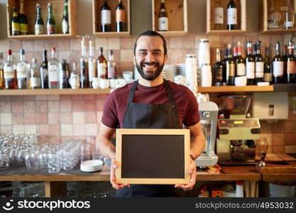 small business, people and service concept - happy man or waiter in apron with black chalkboard banner at bar or coffee shop. happy man or waiter with chalkboard banner at bar