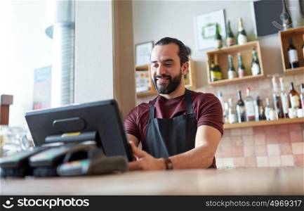 small business, people and service concept - happy man or waiter in apron at counter with cashbox working at bar or coffee shop. happy man or waiter at bar cashbox