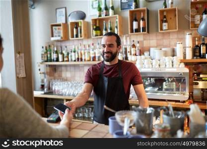 small business, people and service concept - happy man or waiter in apron with card reader and customer with smartphone paying at bar of coffee shop. barman and woman with card reader and smartphone