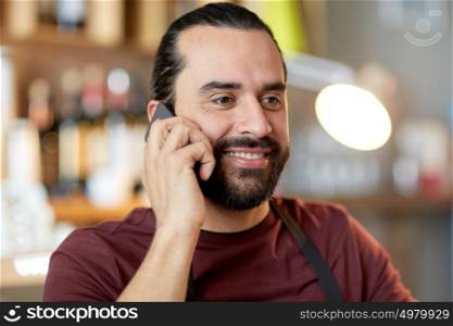 small business, people and service concept - happy man or waiter in apron calling on smartphone at bar. happy man or waiter at bar calling on smartphone