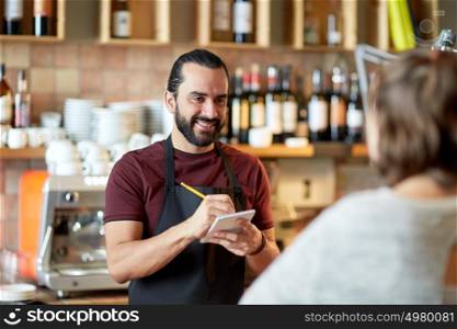 small business, people and service concept - happy man or waiter with notebook and pencil serving customer at coffee shop or bar. man or waiter serving customer at bar