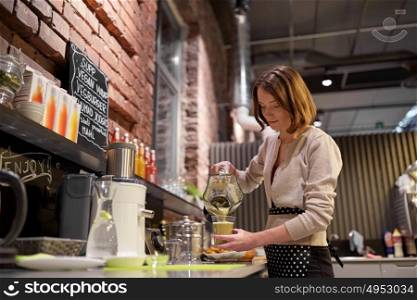 small business, people and service concept - happy woman or barmaid pouring smoothie from blender to cup at vegan cafe. woman or barmaid cooking smoothie at vegan cafe