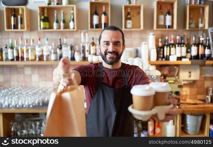 small business, people, takeaway and service concept - happy man or waiter in apron holding coffee cups and paper bag at bar. man or waiter with coffee and paper bag at bar