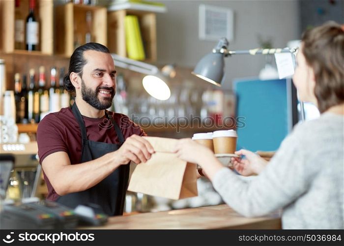small business, people, takeaway and service concept - happy man or waiter giving bag and paper cups with hot drinks to customer at coffee shop. man or waiter serving customer at coffee shop
