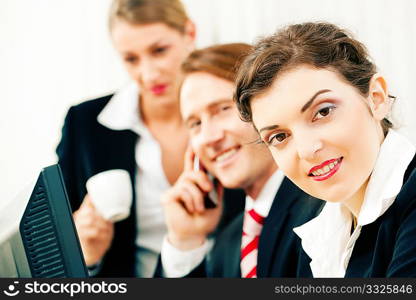 Small business team - man and two women - working in the office using a telephone and looking on a computer screen, one woman drinking a mug of coffee