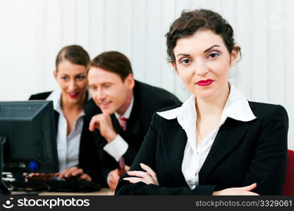 Small business team working in the office on their monitors or laptops in a shared project; one woman is looking in the camera