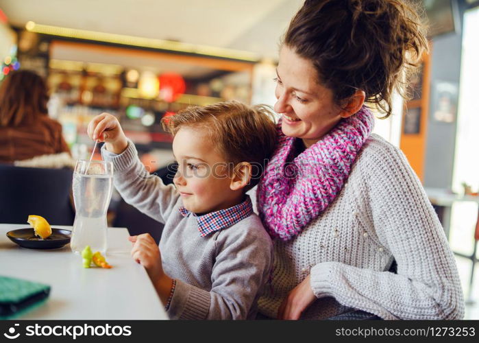Small caucasian boy side view three years old mother and son young woman sitting by the table at the restaurant or cafe child holding plastic spoon or straw while preparing drink having fun