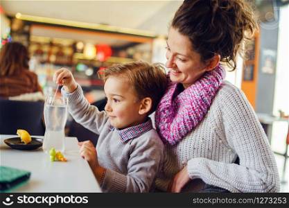 Small caucasian boy side view three years old mother and son young woman sitting by the table at the restaurant or cafe child holding plastic spoon or straw while preparing drink having fun