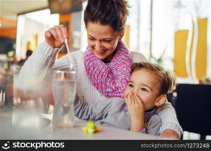 Small caucasian boy three years old mother and son young woman sitting by the table at the restaurant or cafe holding hand over mouth while preparing drink having fun