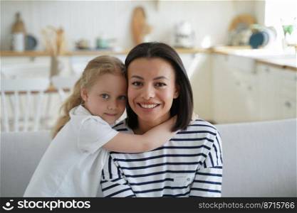 Small child adopted daughter hugs her smiling mother, sitting on sofa at cozy home. Young mom and little kid girl embracing, enjoying time together. Happy motherhood, adoption concept.. Child girl adopted daughter hugs smiling mother, sitting on sofa at home. Happy motherhood, adoption
