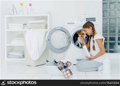 Small child plays with dog russell terrier, poses on knees near washing machine, busy with housekeeping and doing laundry, holds white bottle with washing powder, wears domestic comfortable clothes.