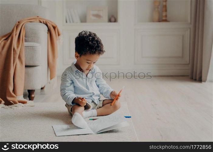Small cute curly mulatto boy sitting on floor in living room in front of colouring book with felt tip pen in hand, child deciding which superhero to colour while spending leisure time at home. Small curly afro mulatto boy with colouring book at home
