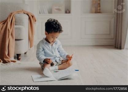 Small cute curly mulatto boy sitting on floor in living room in front of colouring book with felt tip pen in hand, child deciding which superhero to colour while spending leisure time at home. Small curly afro mulatto boy with colouring book at home