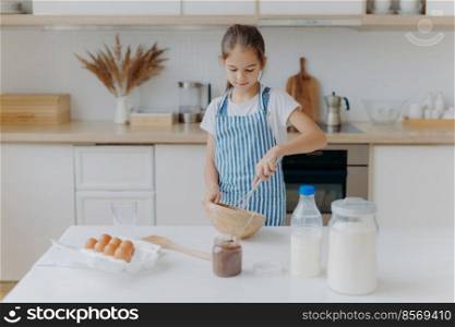 Small cute girl in apron, mixes ingredients, whisks with beater, uses eggs, milk, flour, tries new recipe, stands against kitchen interior, prepares tasty cookies or bakery, learns how to cook.