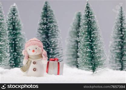 Small cute toy snowman with gift in forest in snow Christmas card design. Snowman with gift in forest