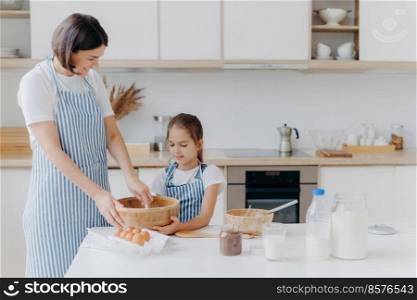 Small daughter and mother prepare cookies together at home, dressed in aprons, stand against kitchen interior, prepare sweet meal together, enjoy spending time at home. Baking, family concept