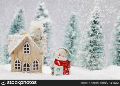 Small decorative snowman near wooden house in fir forest under falling snow. Snowman and house in winter