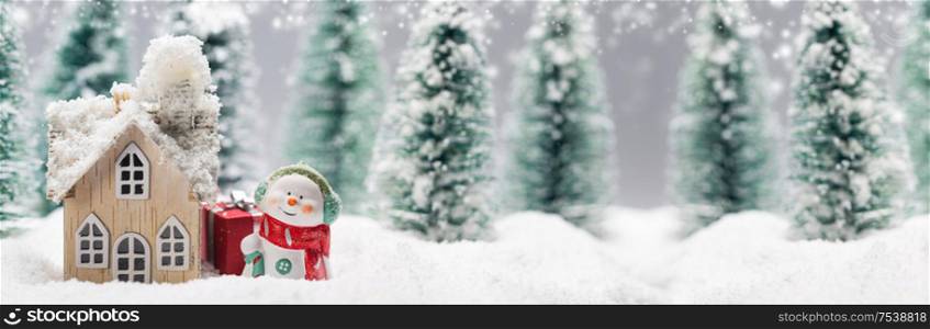 Small decorative snowman with gift near wooden house in fir forest under falling snow. Snowman and house in winter
