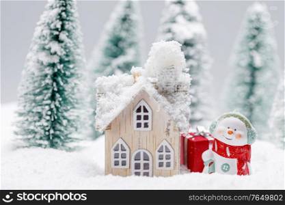 Small decorative snowman with gift near wooden house in fir forest under falling snow. Snowman and house in winter